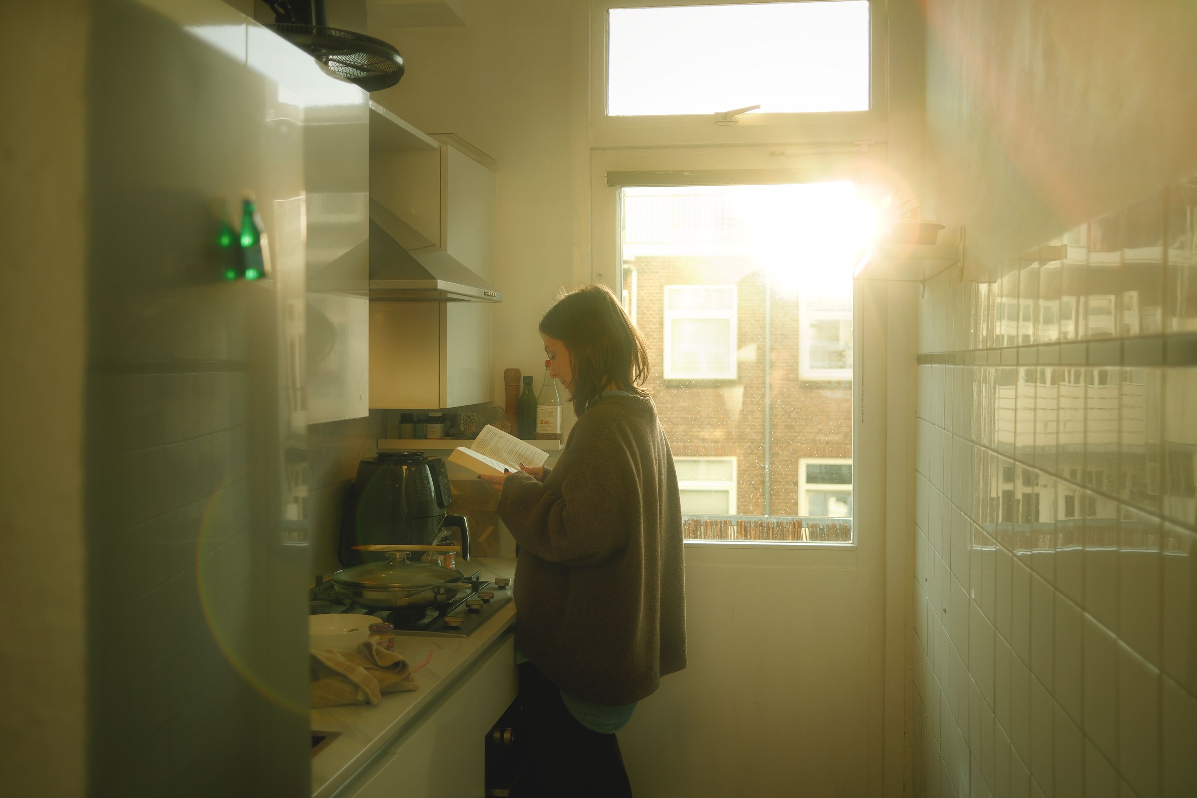 A woman reads a book by the window in a cozy kitchen bathed in warm sunlight
